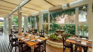 Un área de comedor al aire libre con mesas y sillas de madera, ventanas amplias y una jardinera central con plantas.
