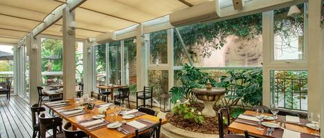 Un área de comedor al aire libre con mesas y sillas de madera, ventanas amplias y una jardinera central con plantas.