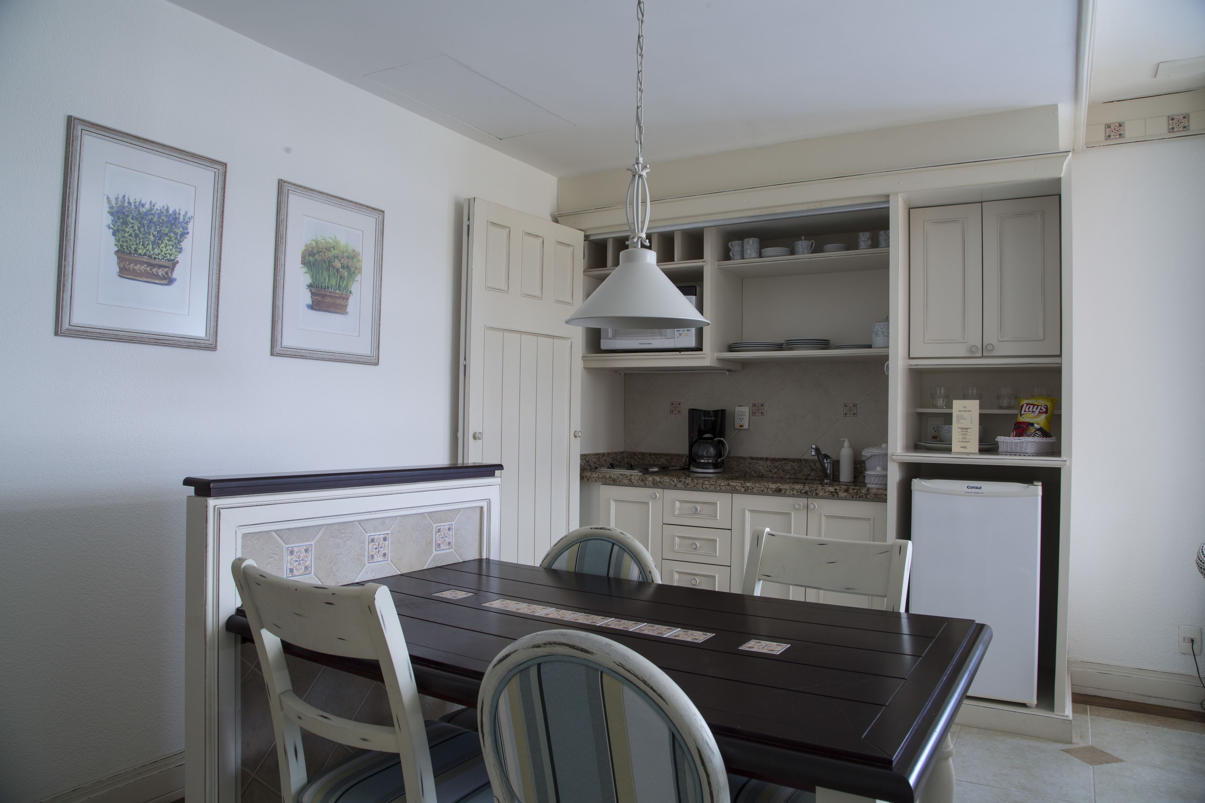 Una cocina con armarios blancos, una mesa de madera oscura y sillas. Hay dos cuadros en la pared, una lámpara en el techo y un refrigerador.