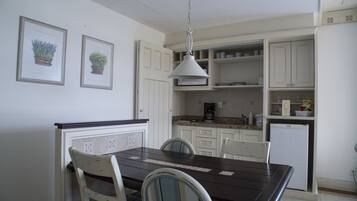 Una cocina con armarios blancos, una mesa de madera oscura y sillas. Hay dos cuadros en la pared, una lámpara en el techo y un refrigerador.