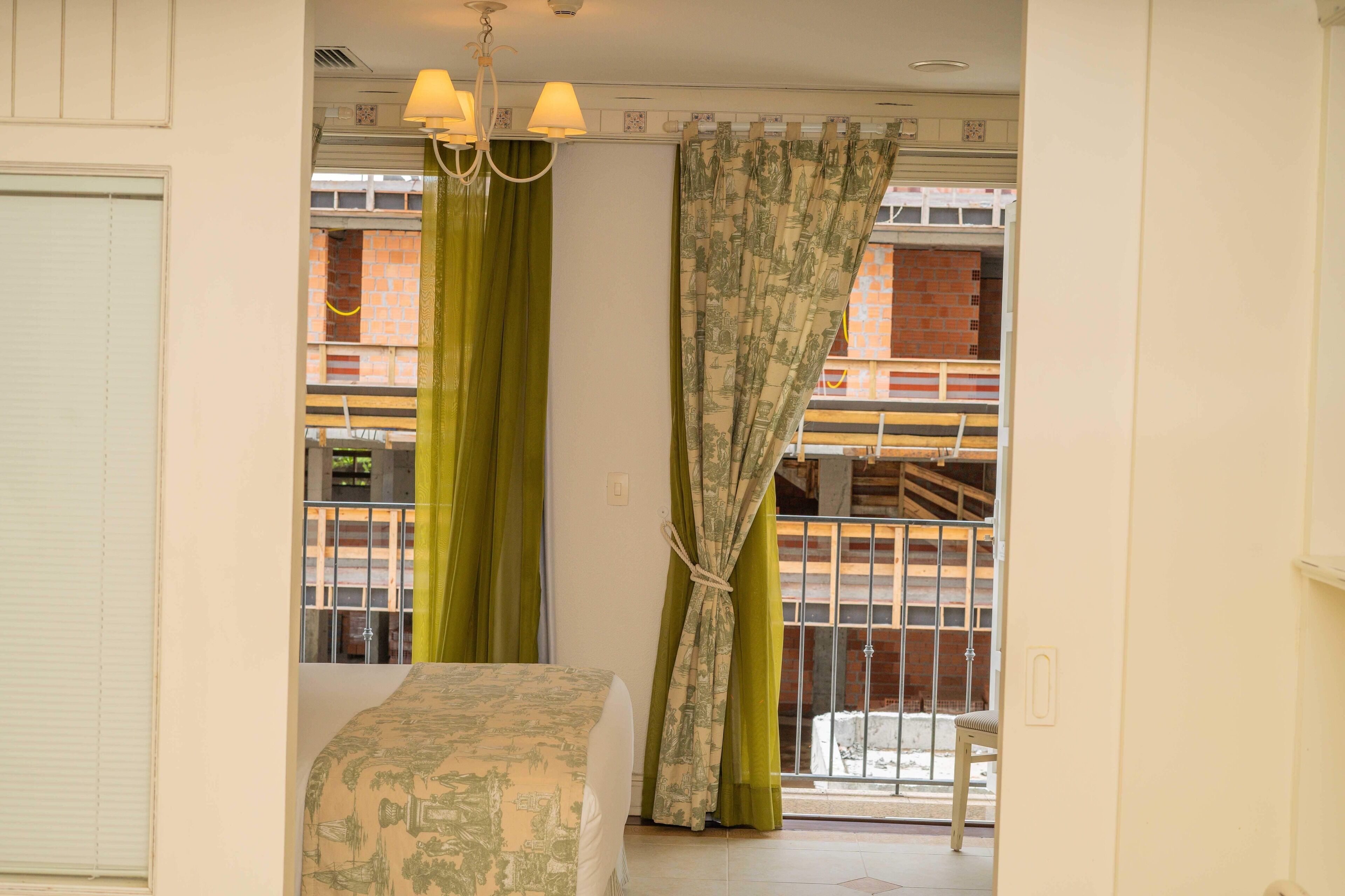 A bedroom with a bed, a window with green curtains, a balcony with a railing, and a chandelier.