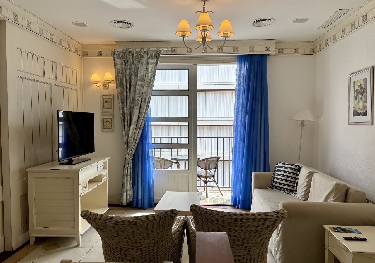 A hotel room with a television, two sofas, a dining table, wicker chairs, a balcony with blue curtains, and a chandelier.