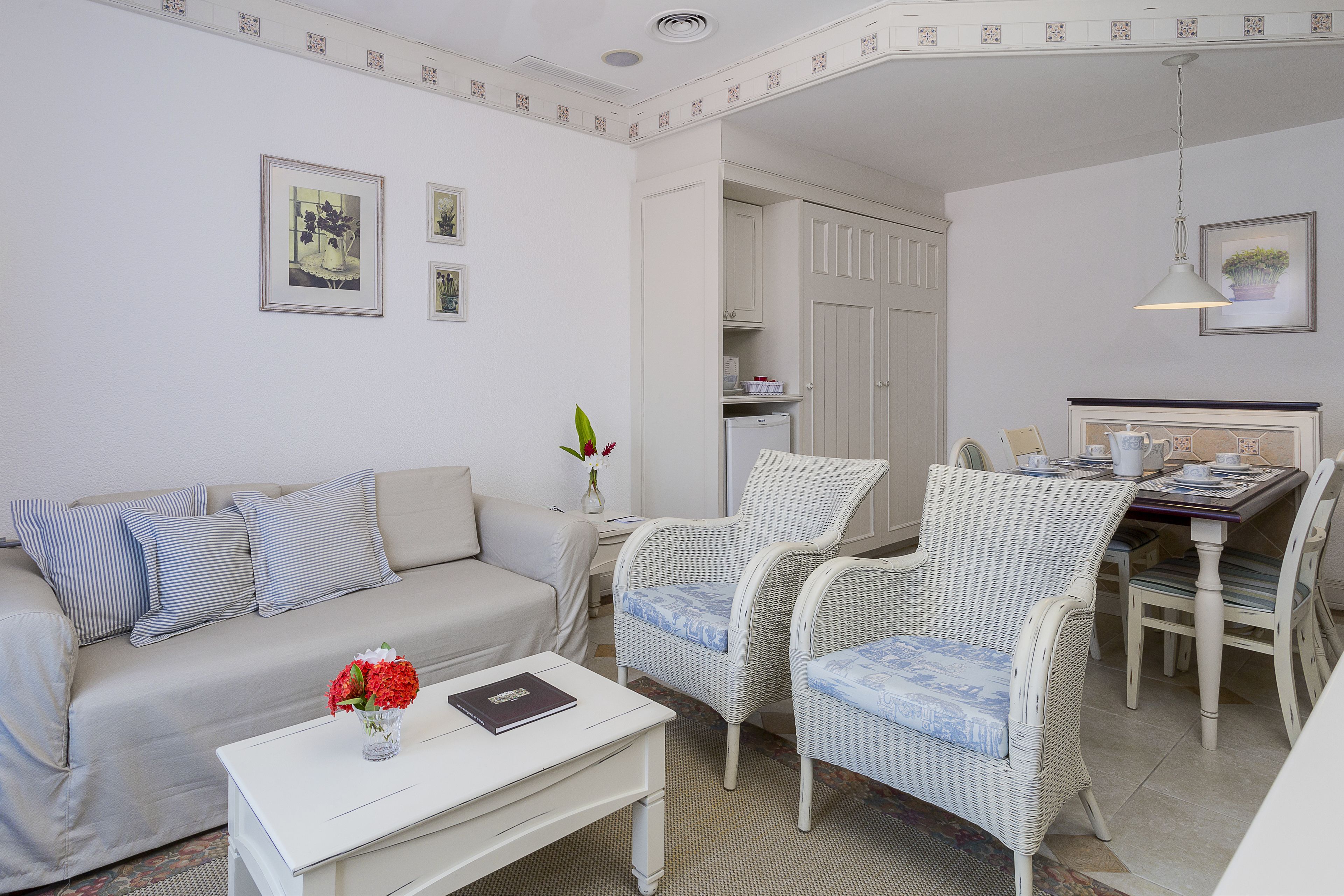 Un salon comprenant un canapé, deux fauteuils en osier, une table basse avec un livre et des fleurs, ainsi qu’un coin repas avec une table et des chaises.