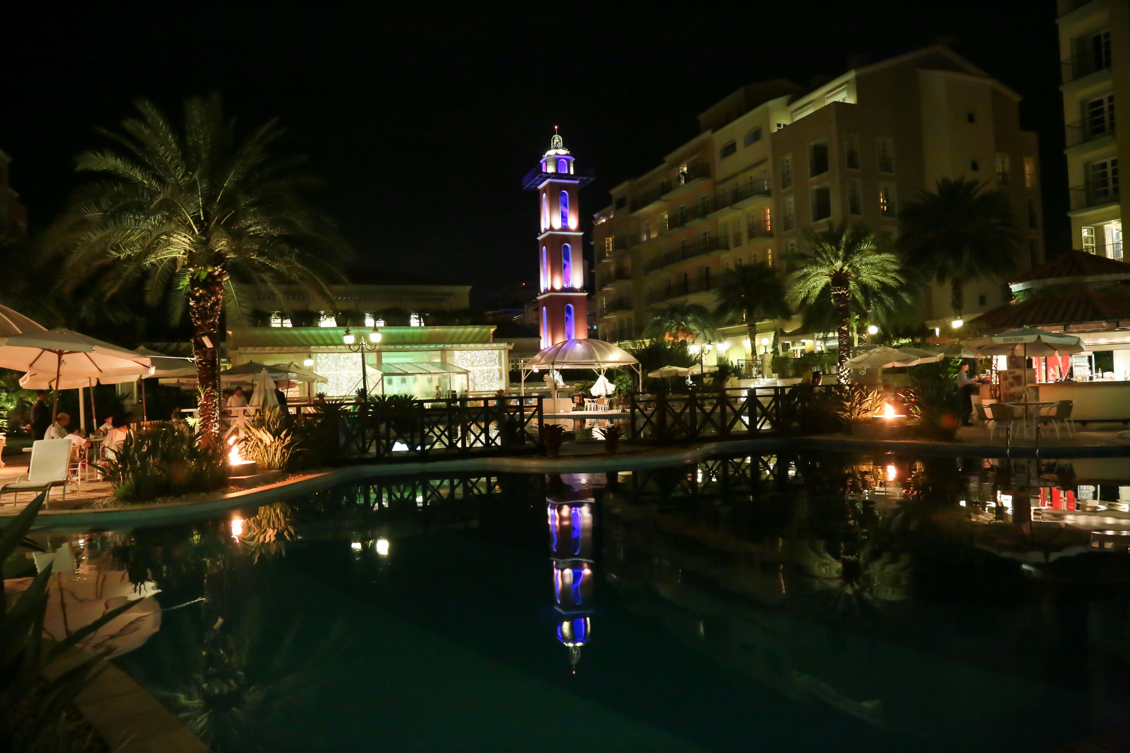 Vue nocturne d’un espace piscine d’hôtel, avec une tour illuminée, des palmiers et des sièges d’extérieur.