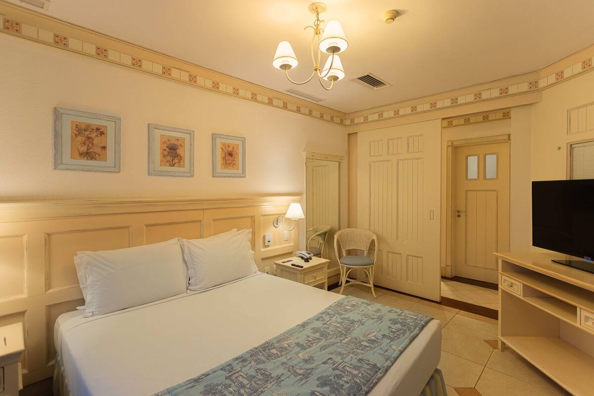 A hotel room with a bed, a chair, a television, and a desk with a lamp.