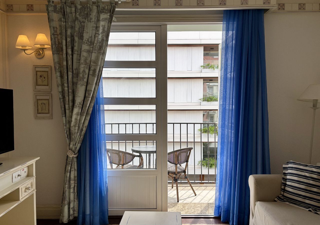 Una habitación con televisión, sofá, balcón con sillas y puerta corredera con cortinas.