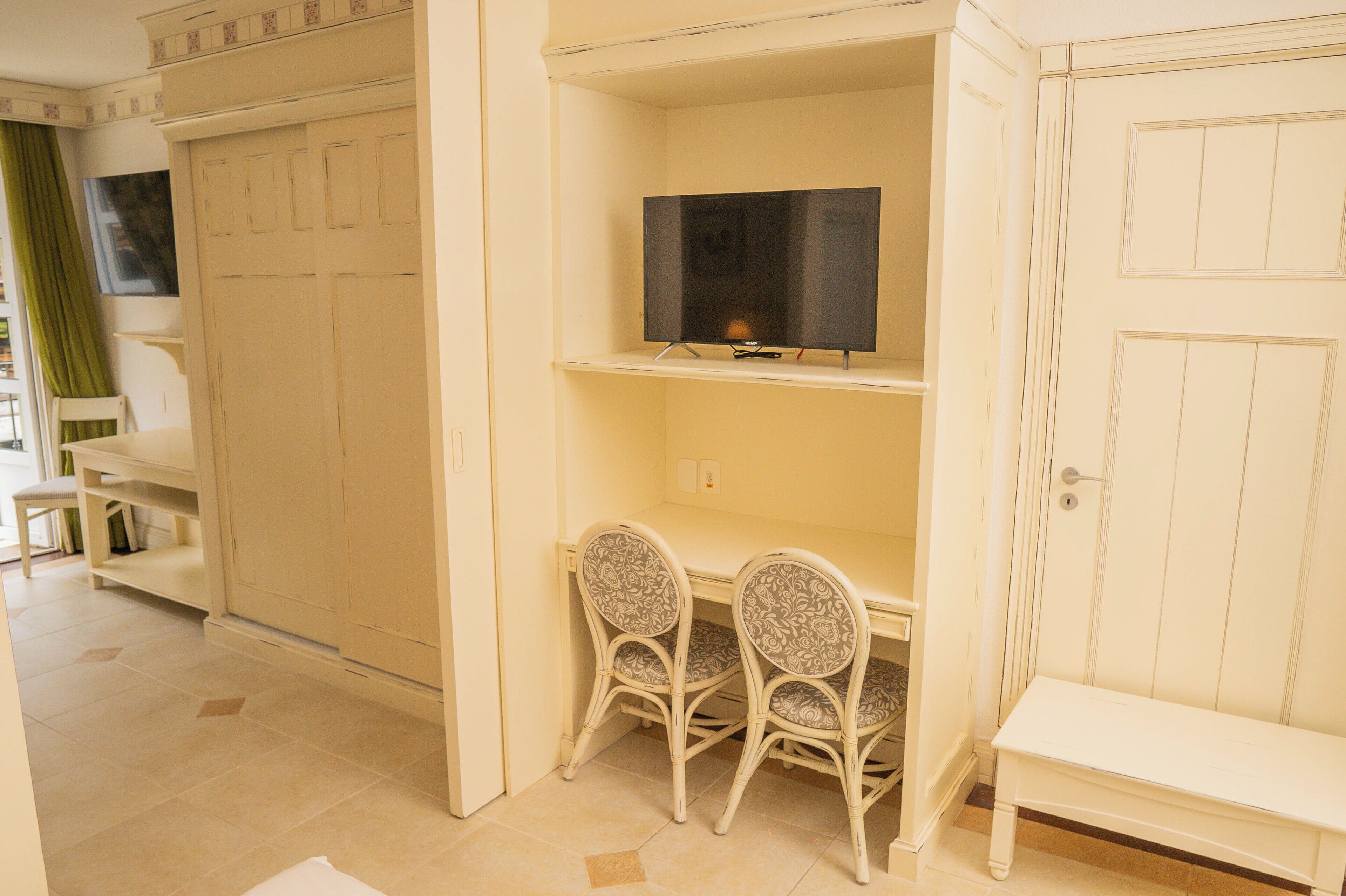 Una habitación con escritorio, dos sillas, televisor en un estante y armario.