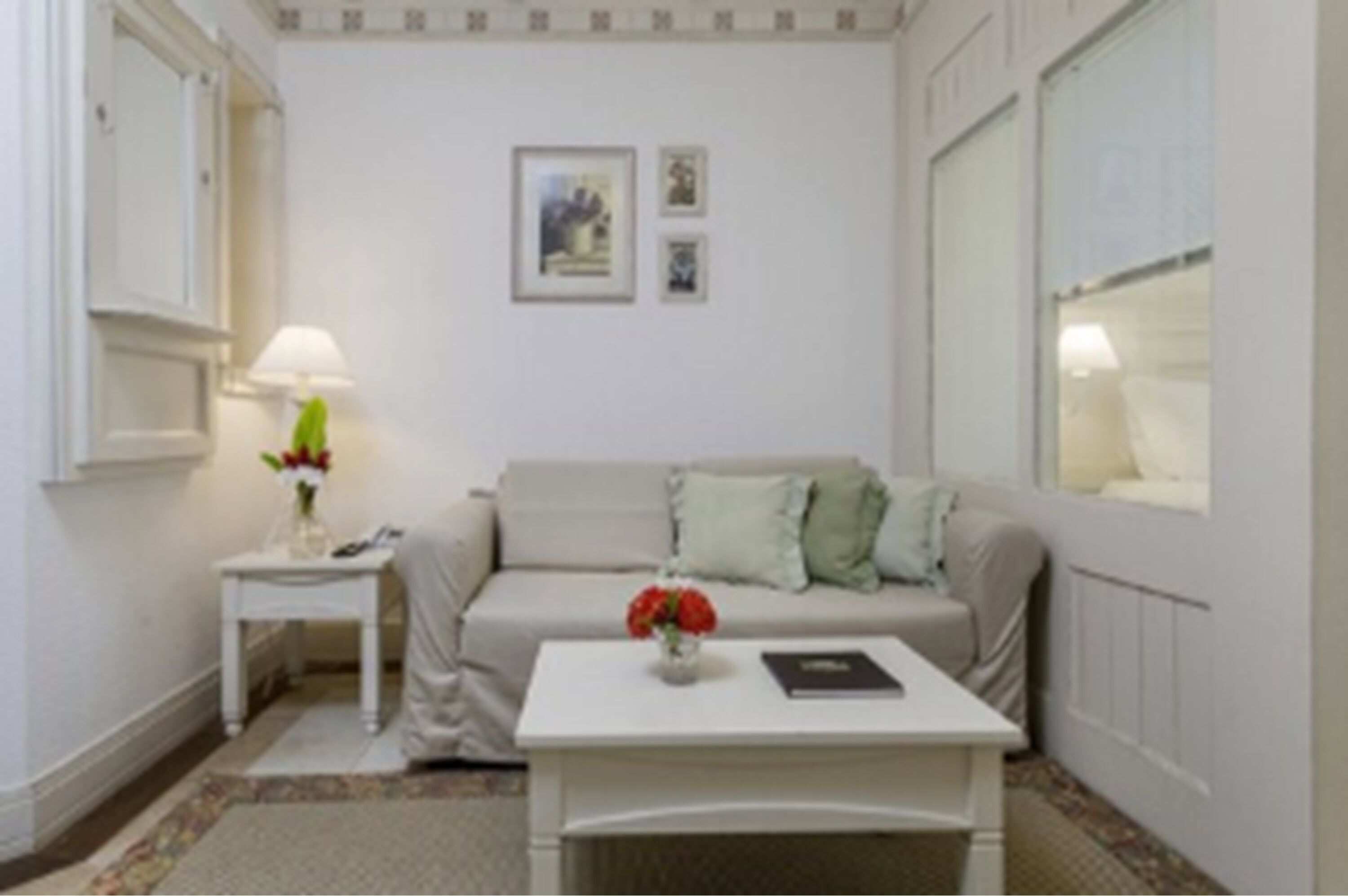 Una habitación pequeña y ordenada con un sofá blanco, una mesa pequeña con un jarrón de flores y una lámpara en una mesita auxiliar.