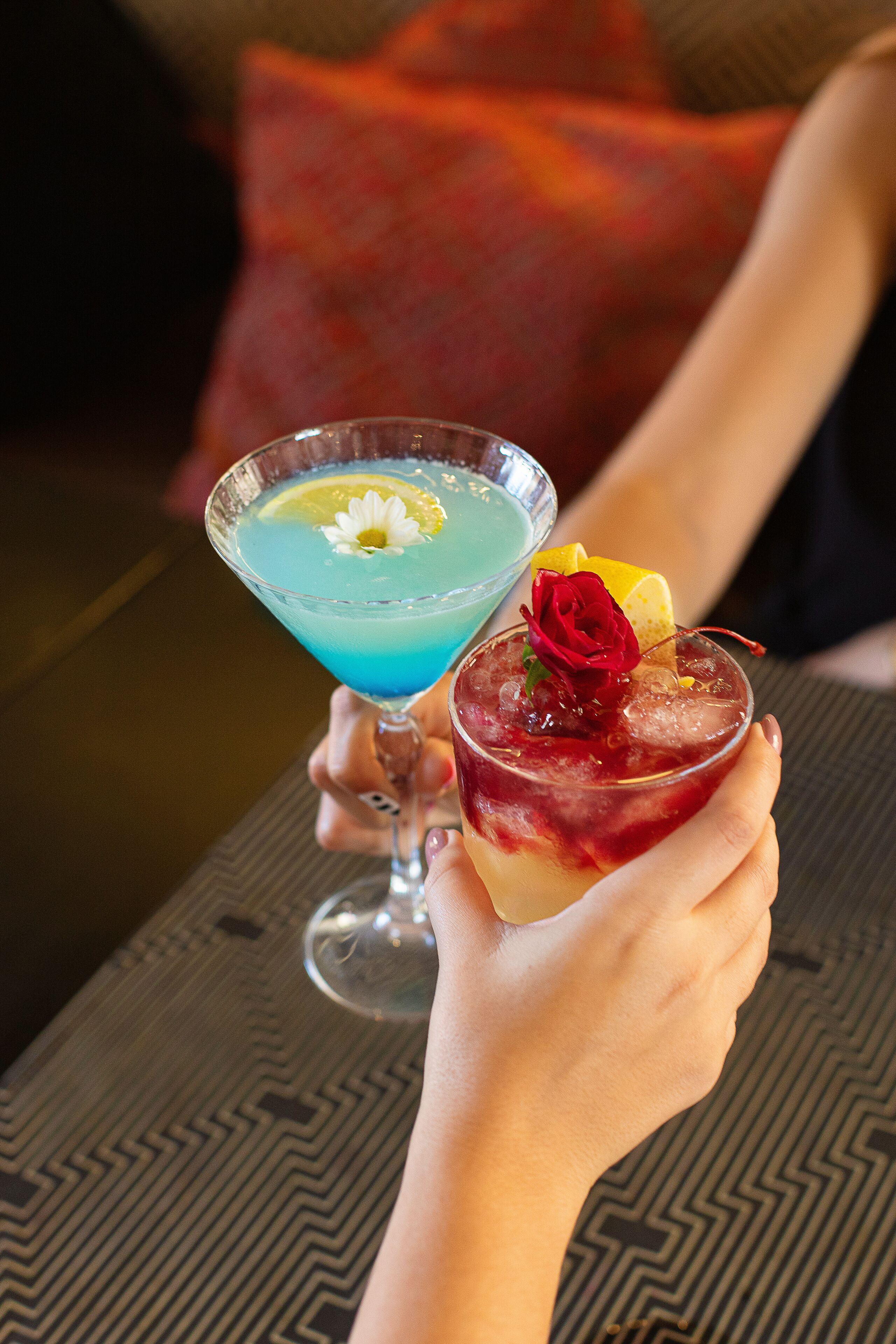 Dos cócteles en copas de martini, uno con una bebida azul y el otro con una bebida roja, ambos adornados con una flor y una rodaja de limón.