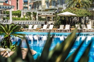 Zona piscina con lettini e ombrelloni, circondata da edifici con balconi e vegetazione.