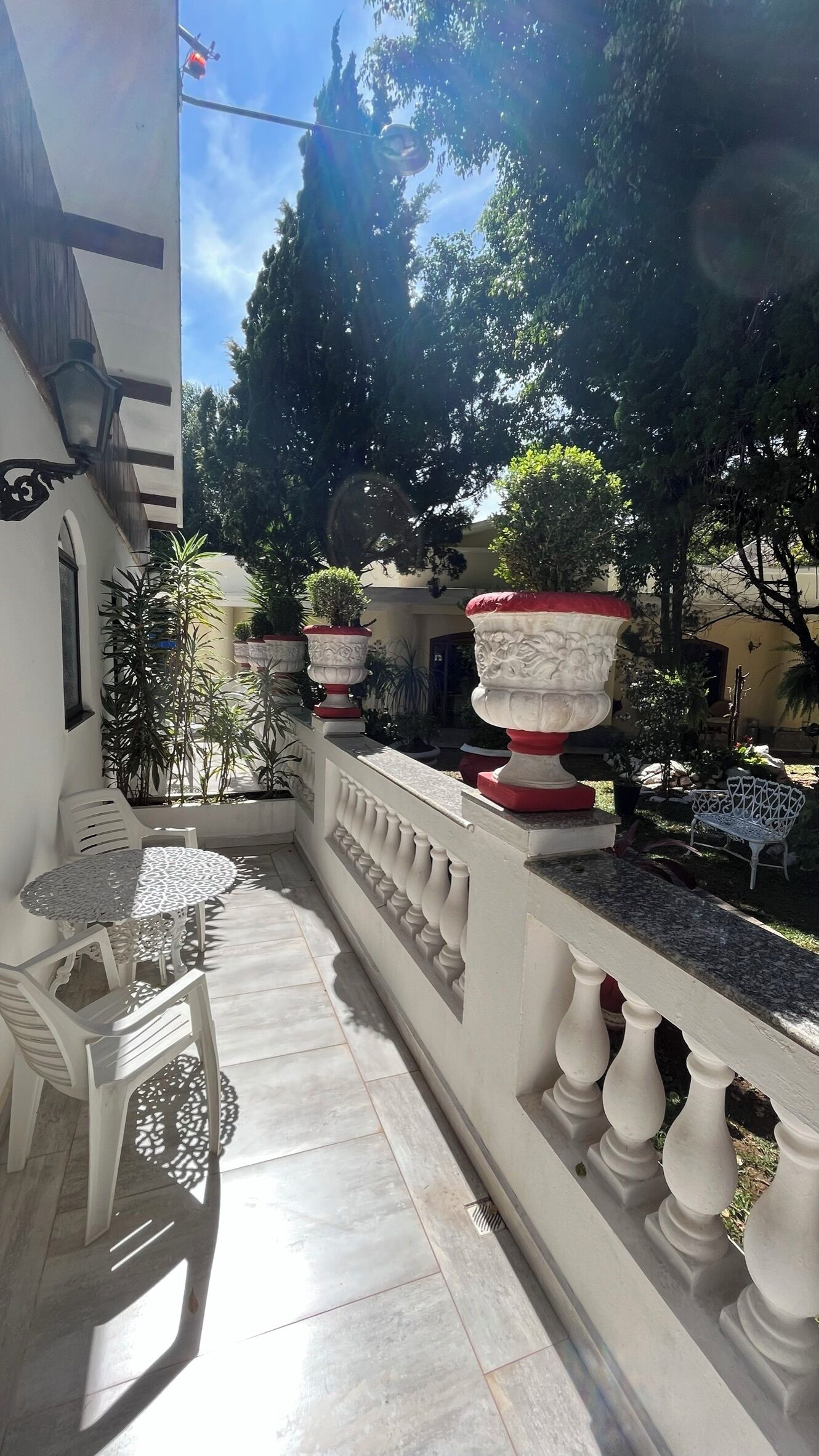 A sunlit outdoor area with white chairs and tables, surrounded by potted plants and a white balustrade.