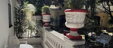 A sunlit outdoor area with white chairs and tables, surrounded by potted plants and a white balustrade.