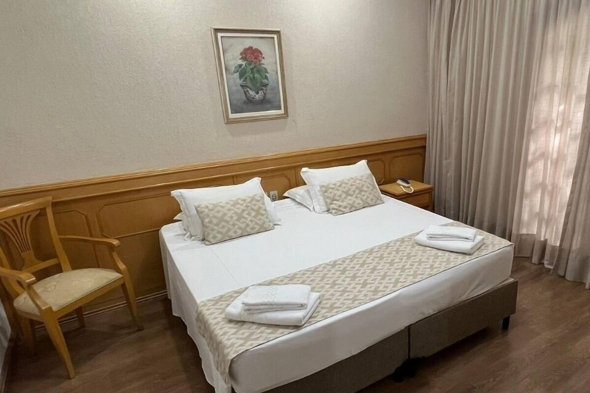 Una cama bien hecha con dos almohadas, una mesita de noche con un teléfono y una silla.