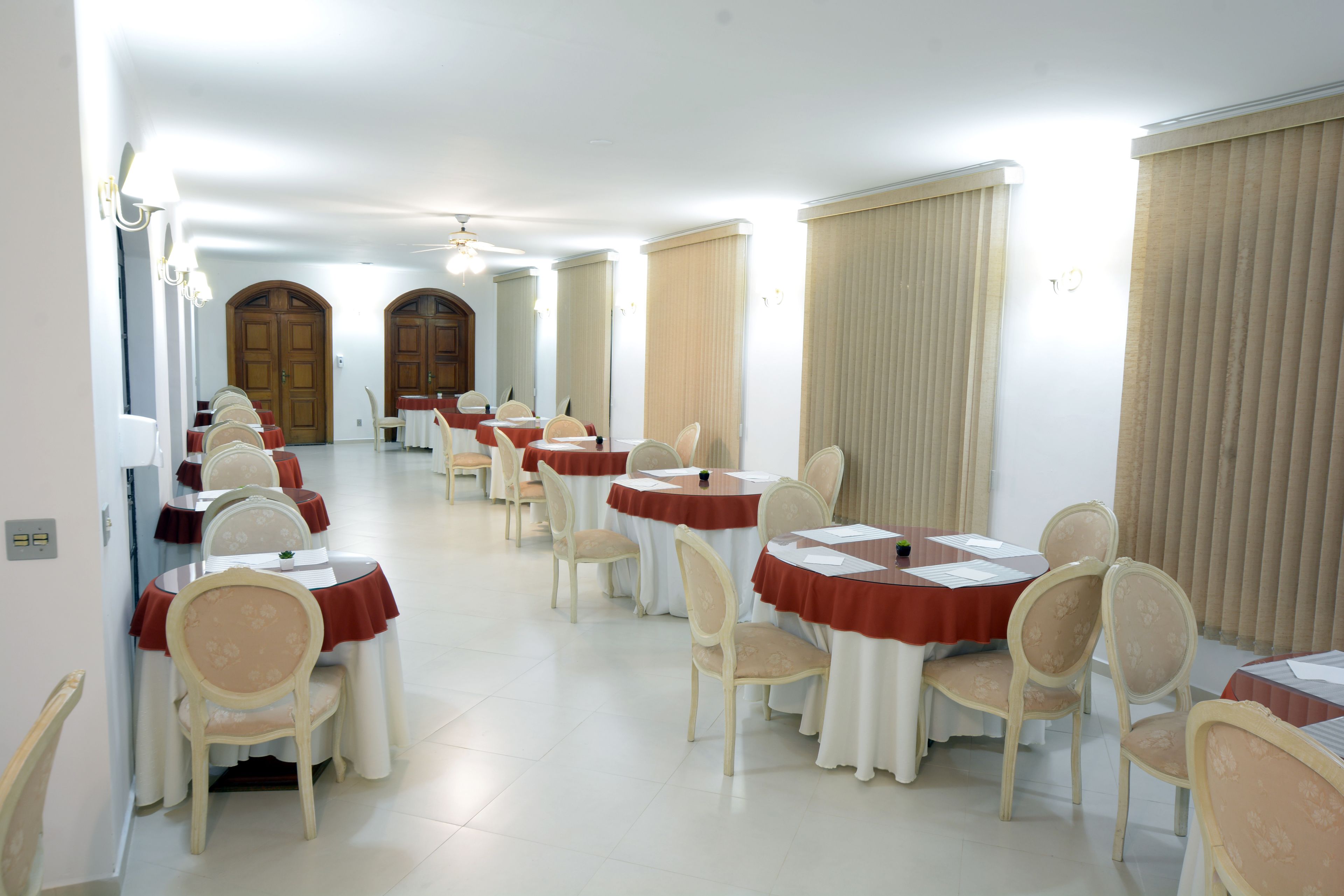 Uma sala de jantar de hotel com várias mesas e cadeiras, dispostas em linha, com teto alto e grandes janelas.
