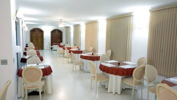 Uma sala de jantar de hotel com várias mesas e cadeiras, dispostas em linha, com teto alto e grandes janelas.