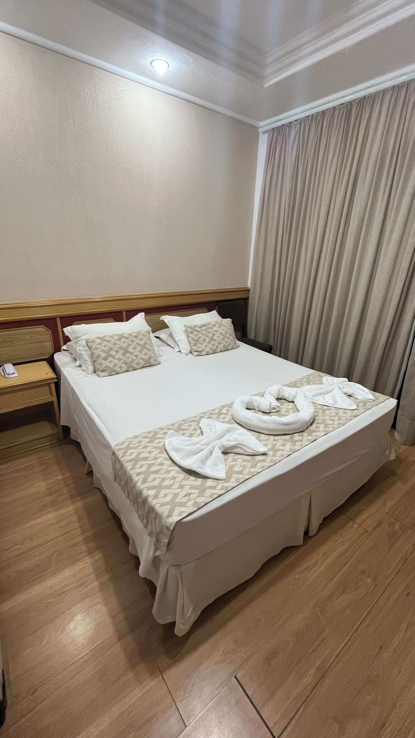 A neatly made bed with white linens and a towel arrangement, a wooden nightstand with a phone, and a wooden floor.