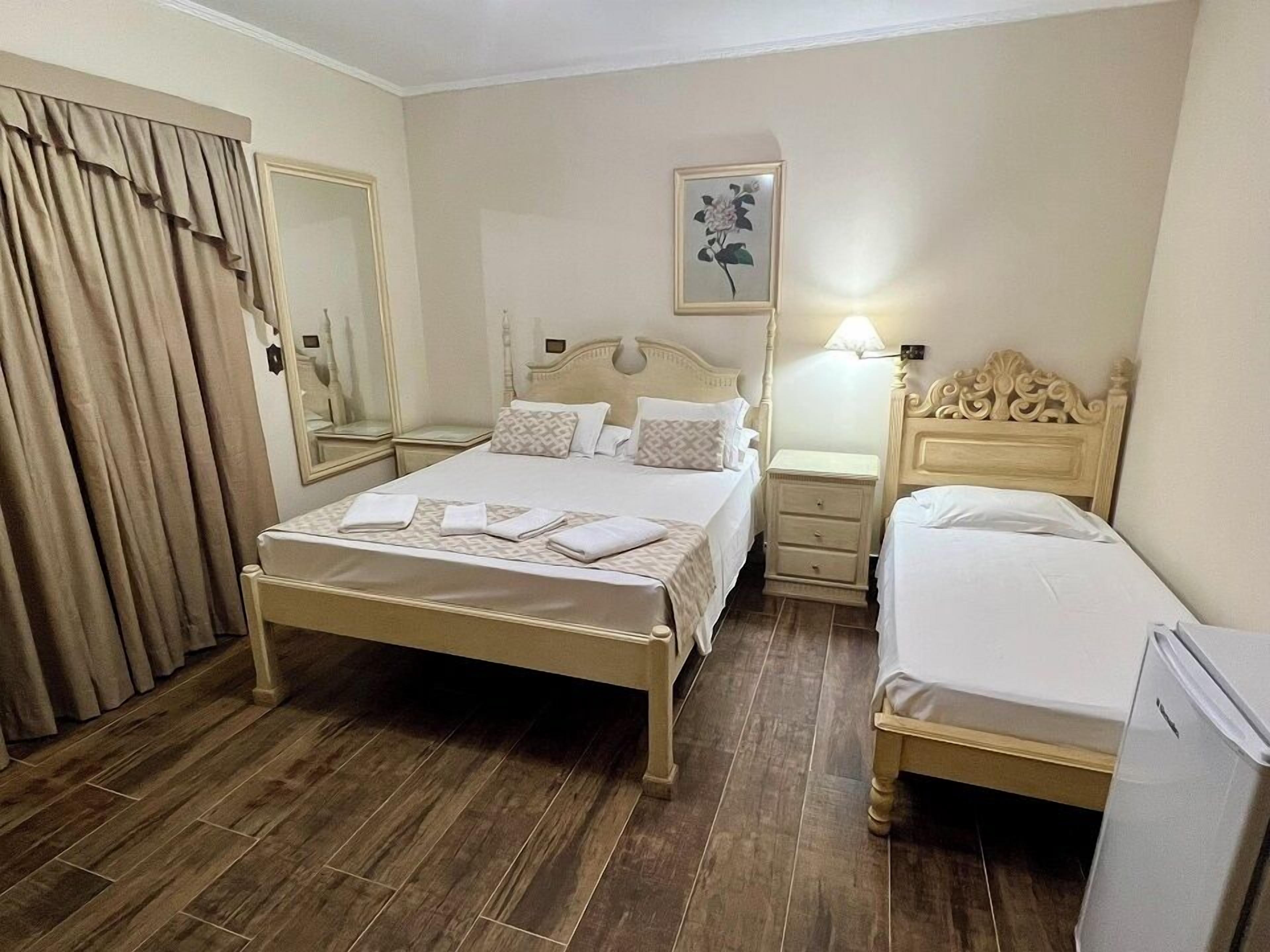 Une chambre d’hôtel avec un grand lit, un lit plus petit, une table de chevet, une commode, un miroir et un tableau au mur.