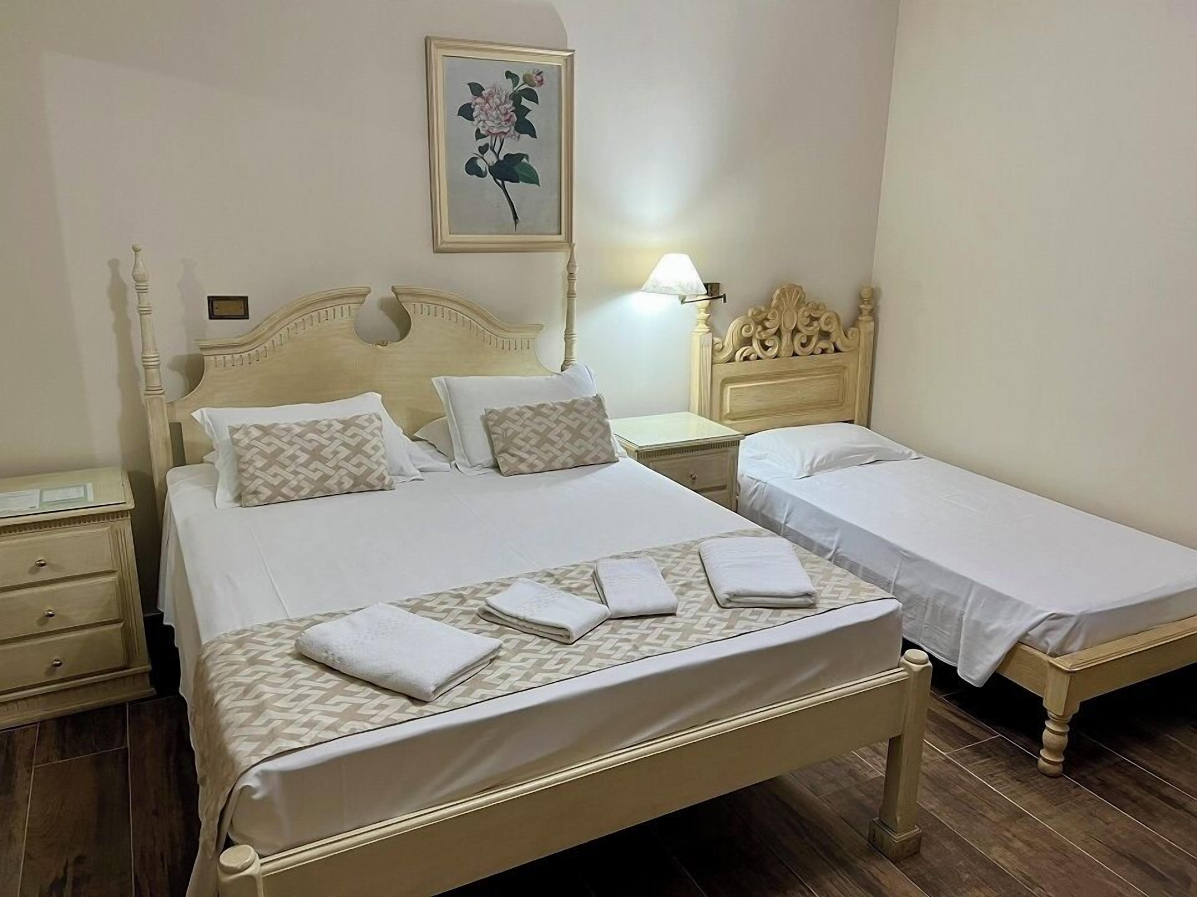 A hotel room with two beds, a nightstand, a lamp, and a framed floral artwork on the wall.