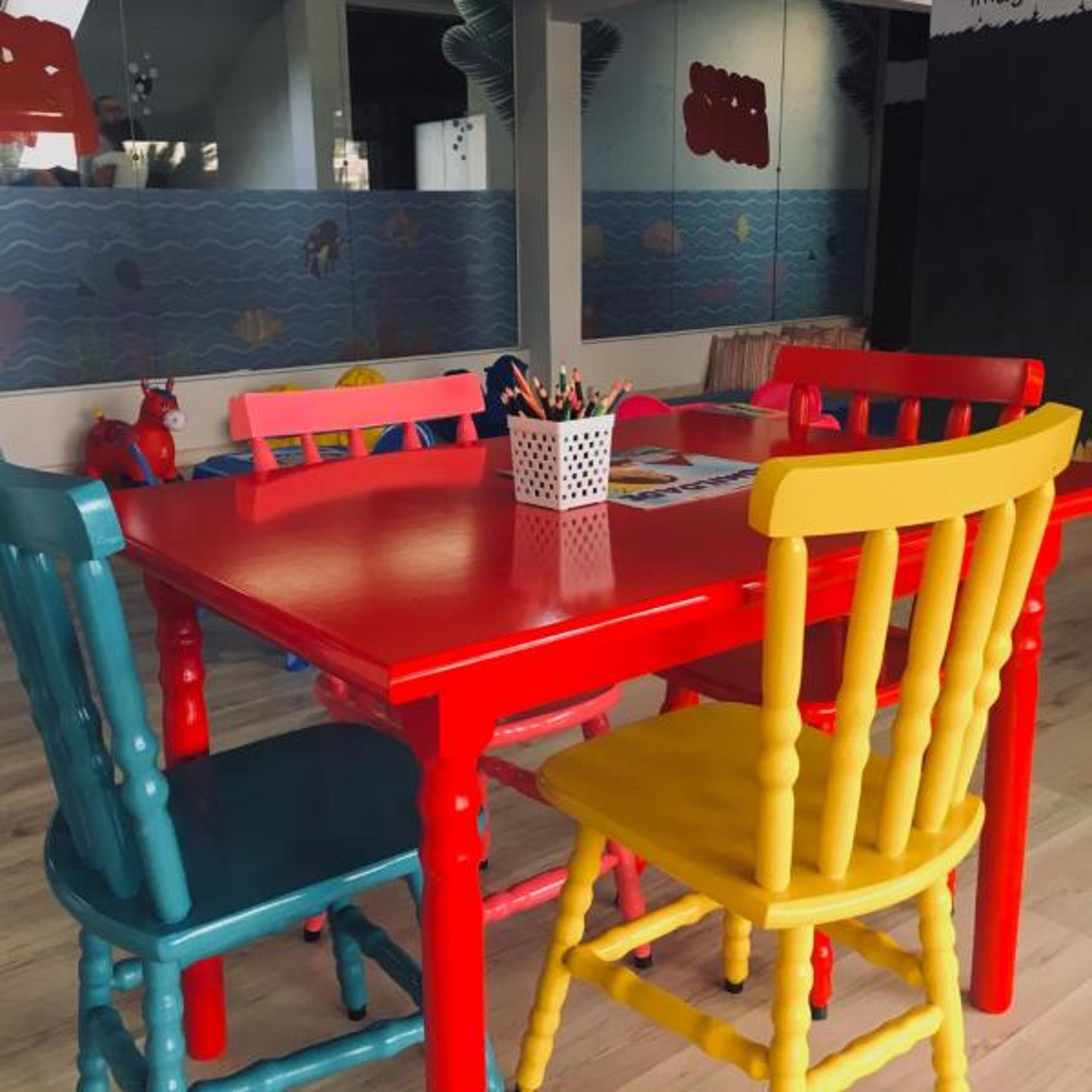 Uma área de refeições com mesa e cadeiras vermelhas, um cesto de lápis e uma parede com um mural.