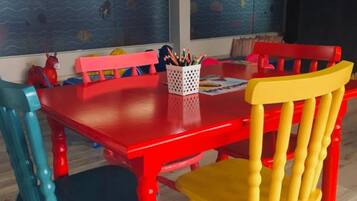Uma área de refeições com mesa e cadeiras vermelhas, um cesto de lápis e uma parede com um mural.