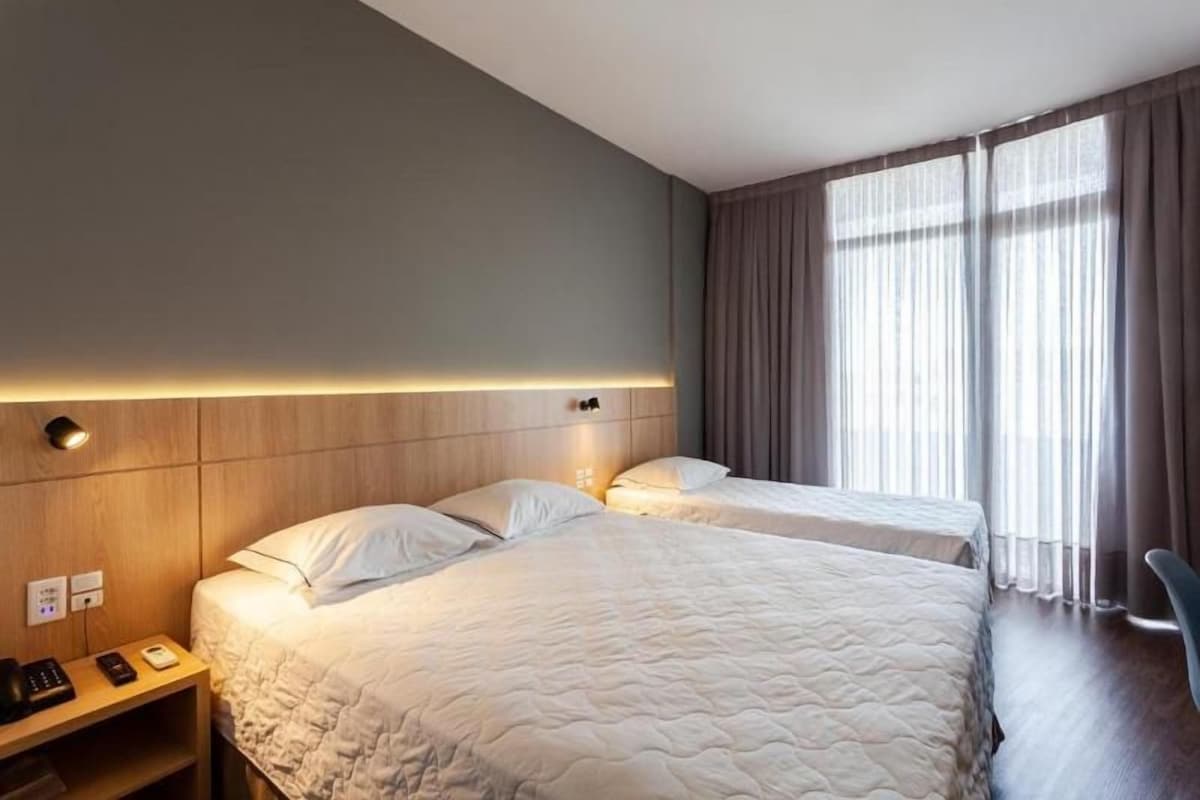 A hotel room with two beds, a bedside table with a lamp, a chair, and a large window with curtains.