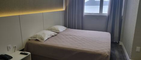 A hotel room with a bed, a bedside table with a phone, and a window with curtains.