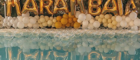 A pool area with gold and white balloons spelling out "NARAIBA" and a reflection of the balloons and pool in the water.