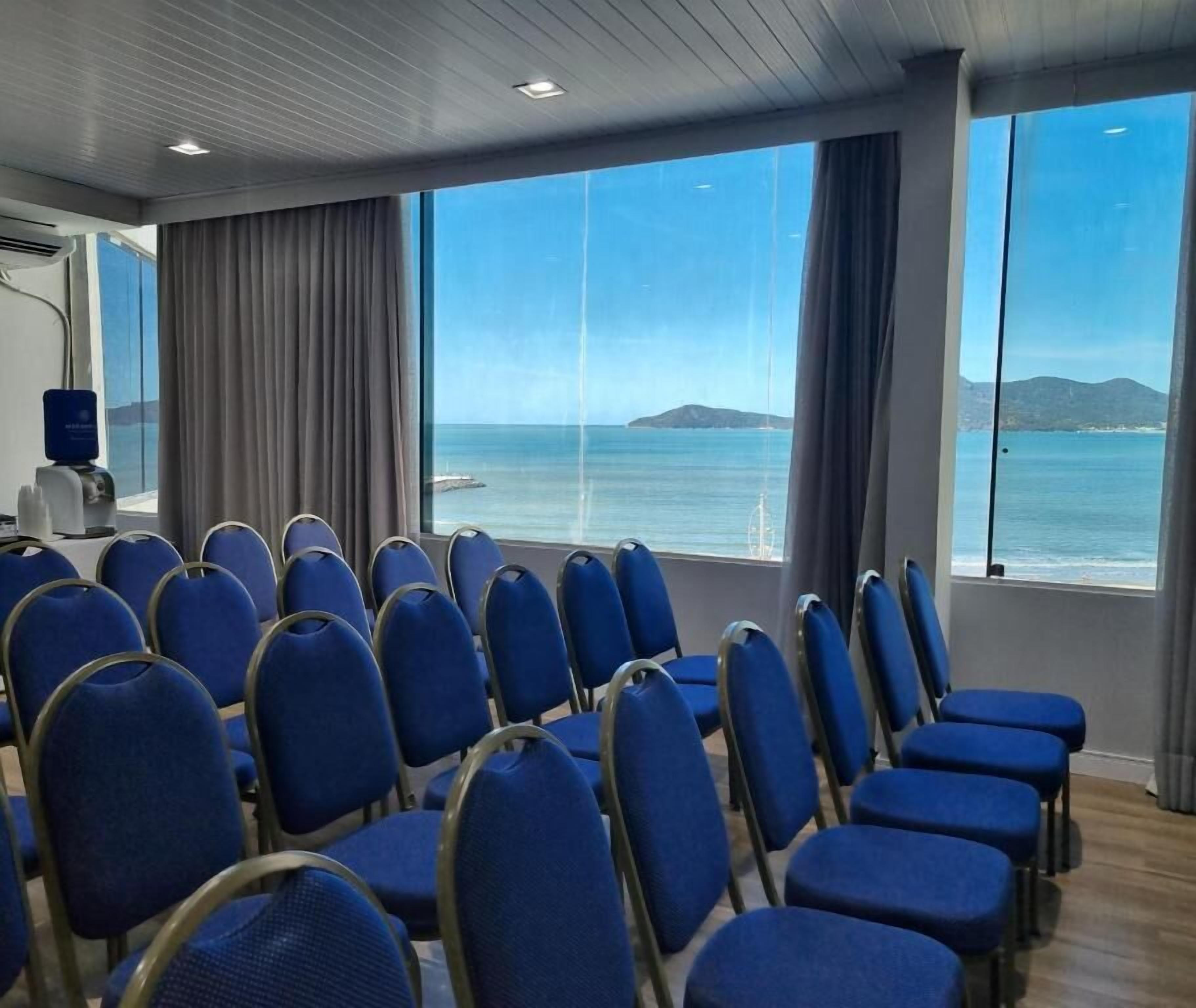 A conference room with rows of blue chairs facing a large window overlooking a body of water and distant land.