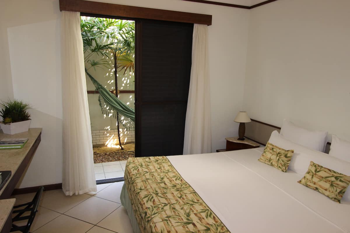 Quarto de hotel com cama, escrivaninha, mesa de cabeceira, luminária e vista para um jardim através de uma porta de correr.