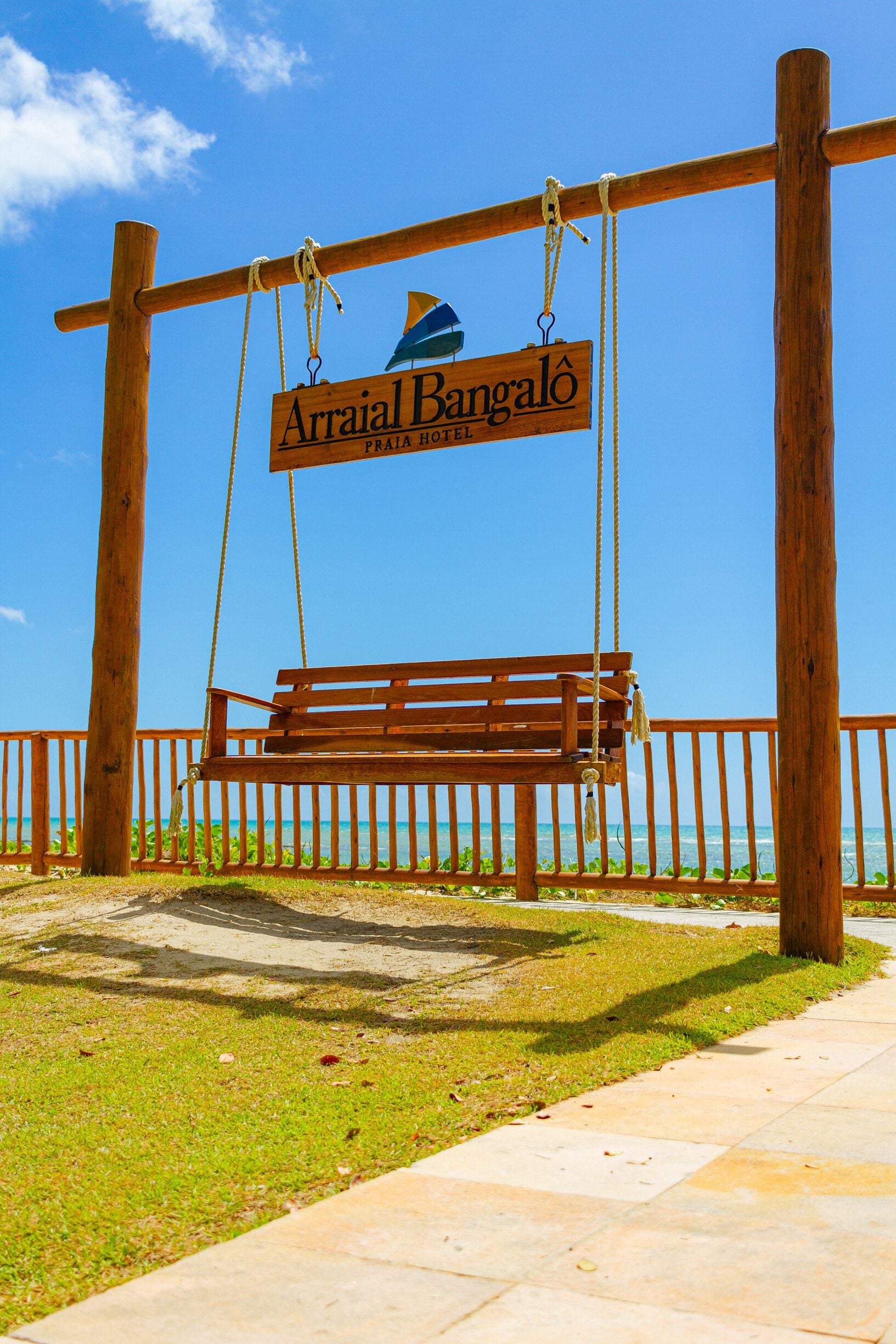 A wooden swing bench under a sign for Arraial Bangalo, with a clear view of the ocean in the background.