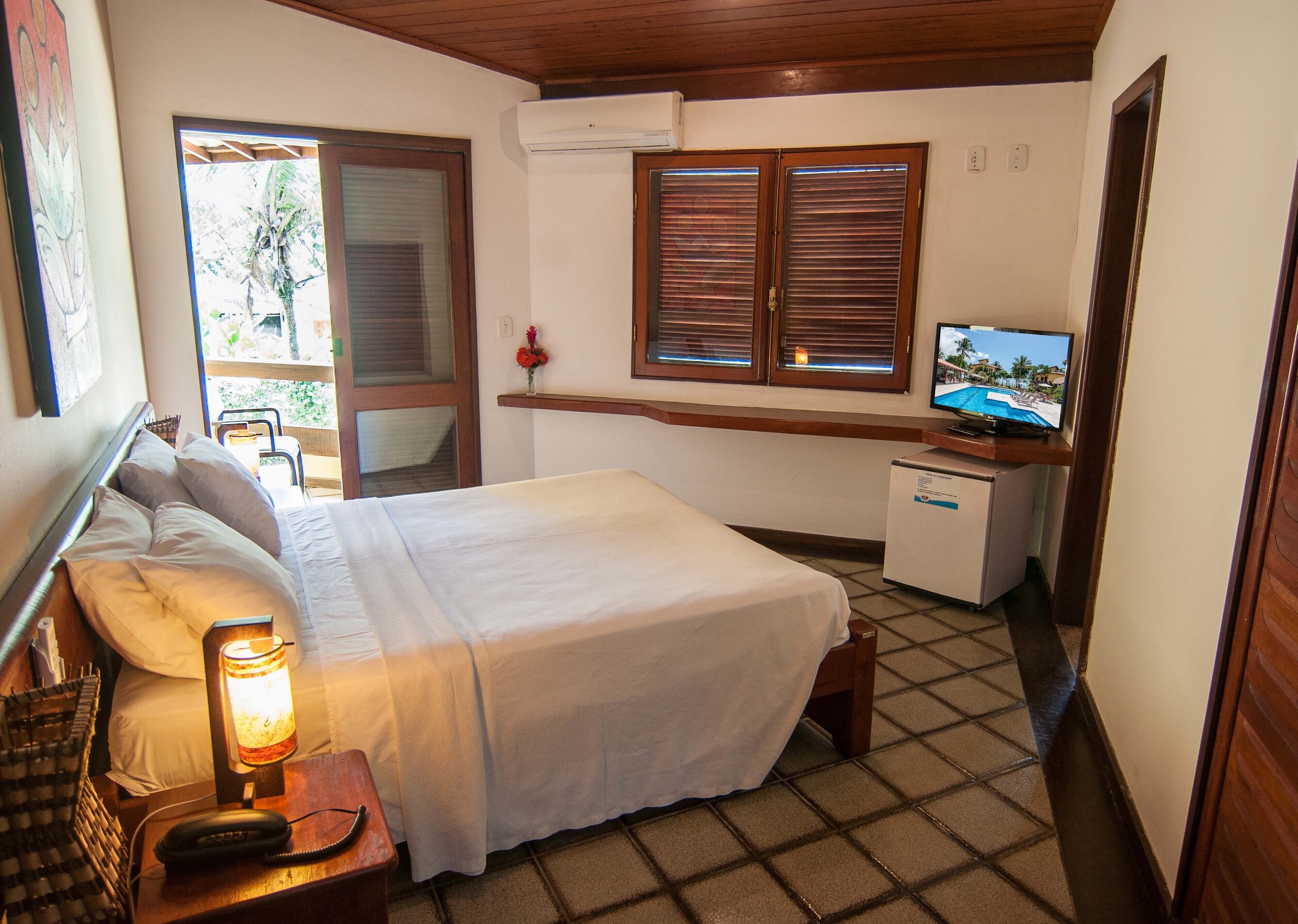 A bedroom with a bed, bedside lamp, a desk with a computer, a TV, and a window with blinds.