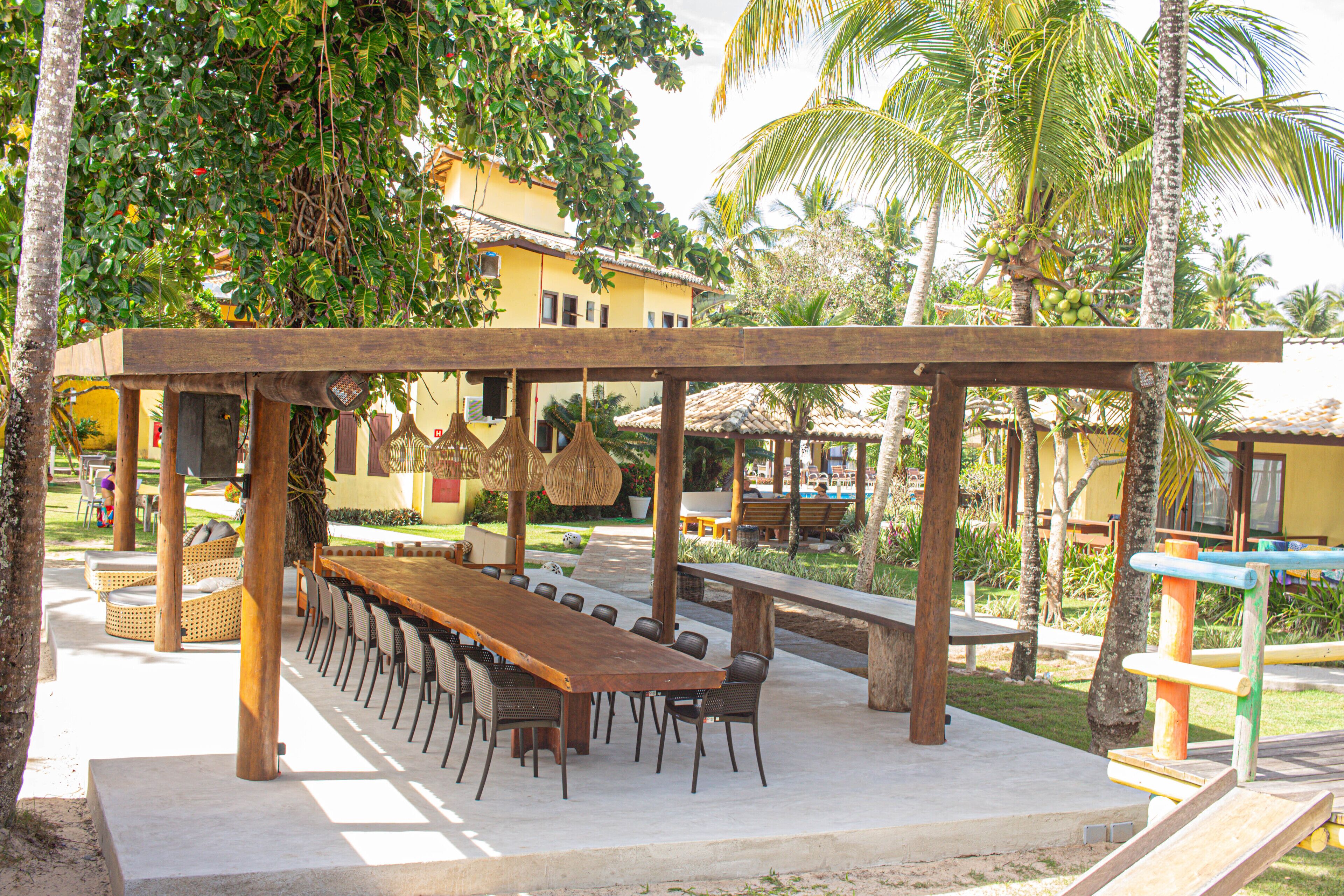 Un espace de restauration en plein air avec une longue table et des chaises en bois, entouré de palmiers et d’un bâtiment jaune en arrière-plan.