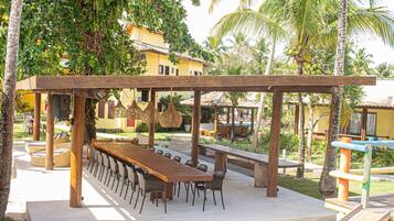 Un espace de restauration en plein air avec une longue table et des chaises en bois, entouré de palmiers et d’un bâtiment jaune en arrière-plan.