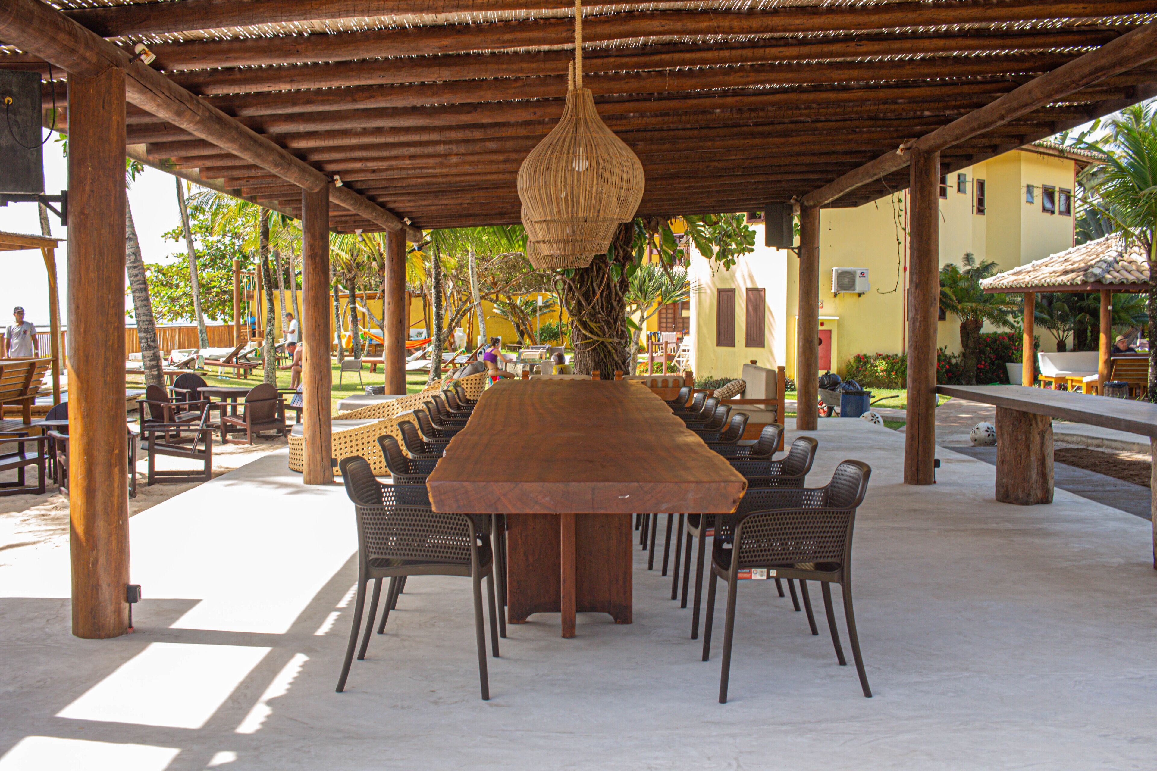 A covered outdoor dining area with a long wooden table and chairs, surrounded by wooden pillars and a hanging light fixture.
