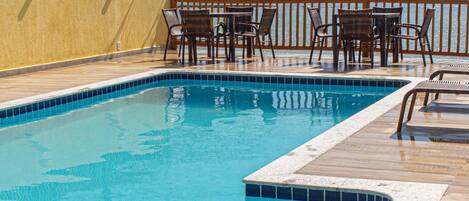 A swimming pool with a wooden deck, surrounded by wicker chairs and tables, overlooking a body of water.