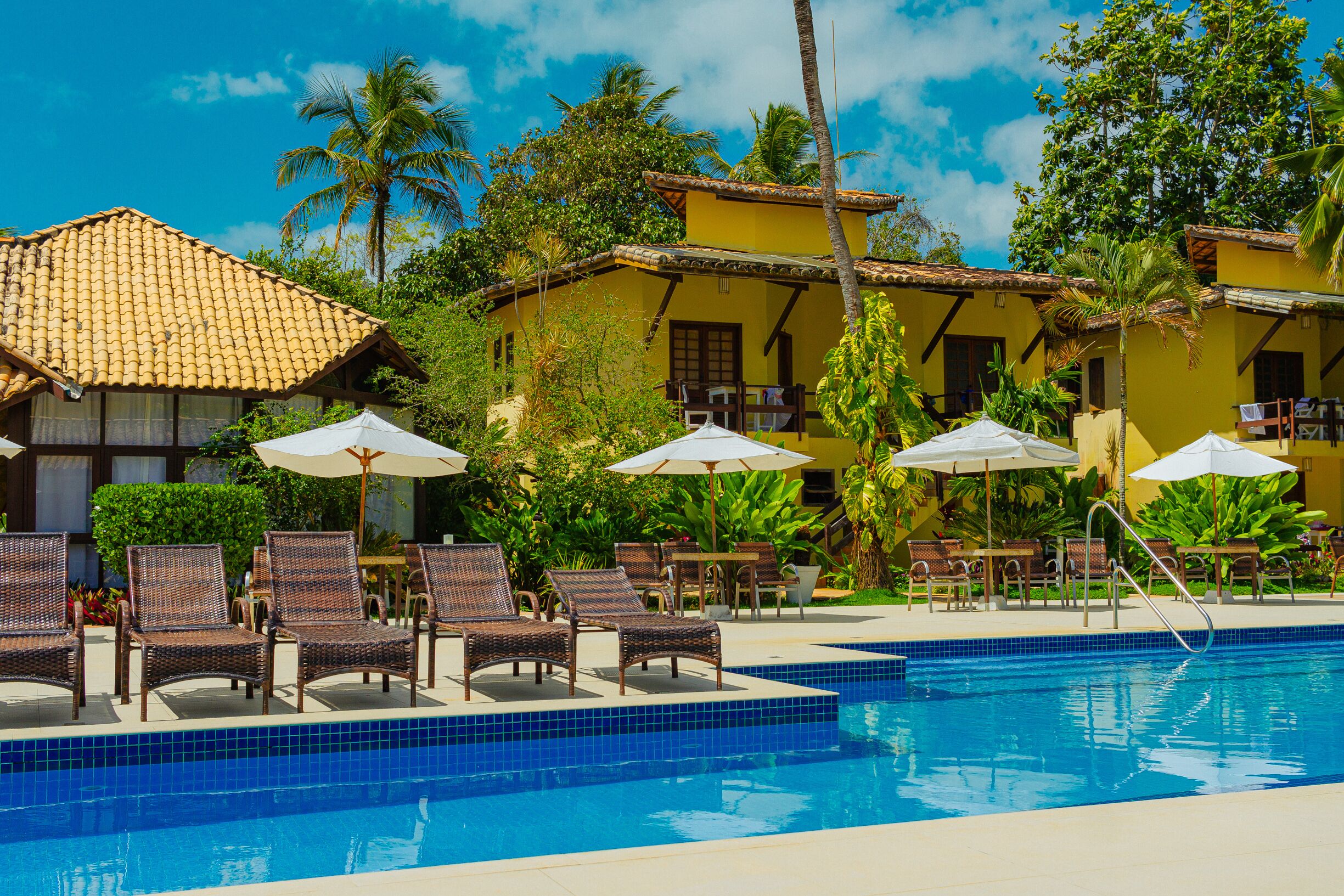 Un área de piscina con sillones de descanso y sombrillas, rodeada de edificios amarillos y palmeras.