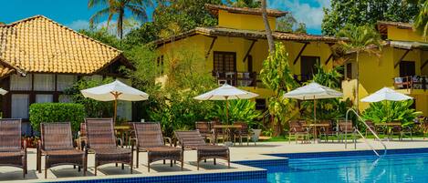 Un área de piscina con sillones de descanso y sombrillas, rodeada de edificios amarillos y palmeras.