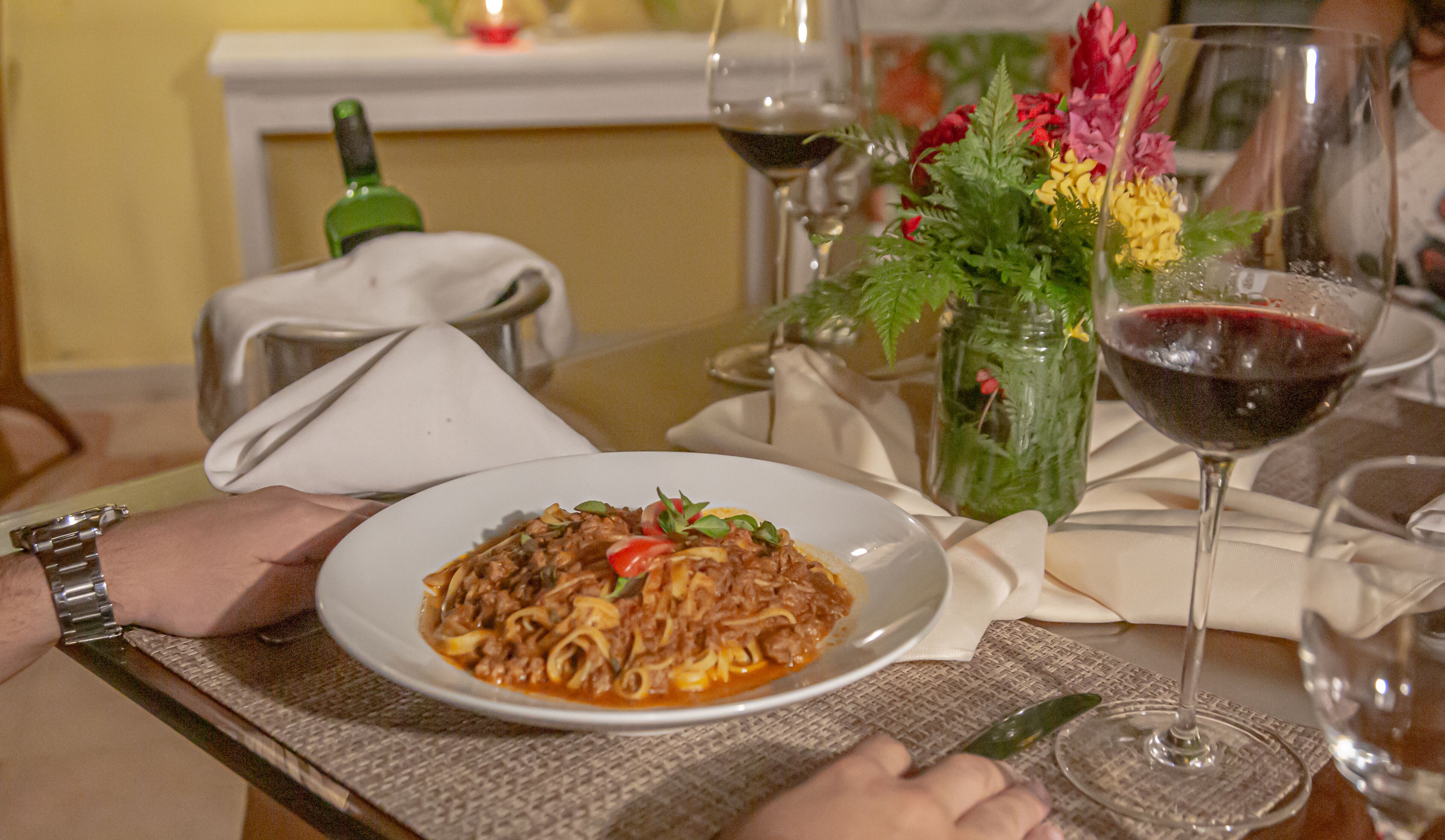 Una mesa de comedor con un plato de pasta, una copa de vino, una botella verde y un florero con flores.