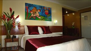 A hotel room with a large bed, a nightstand with a phone, a painting on the wall, and a vase with red flowers. - Atlantida Park Hotel (Porto Seguro)