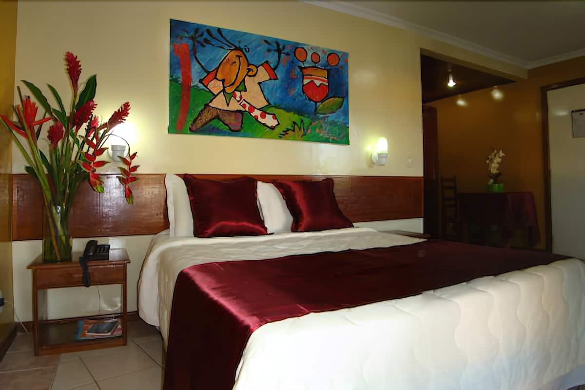 A hotel room with a large bed, a nightstand with a phone, a painting on the wall, and a vase with red flowers.