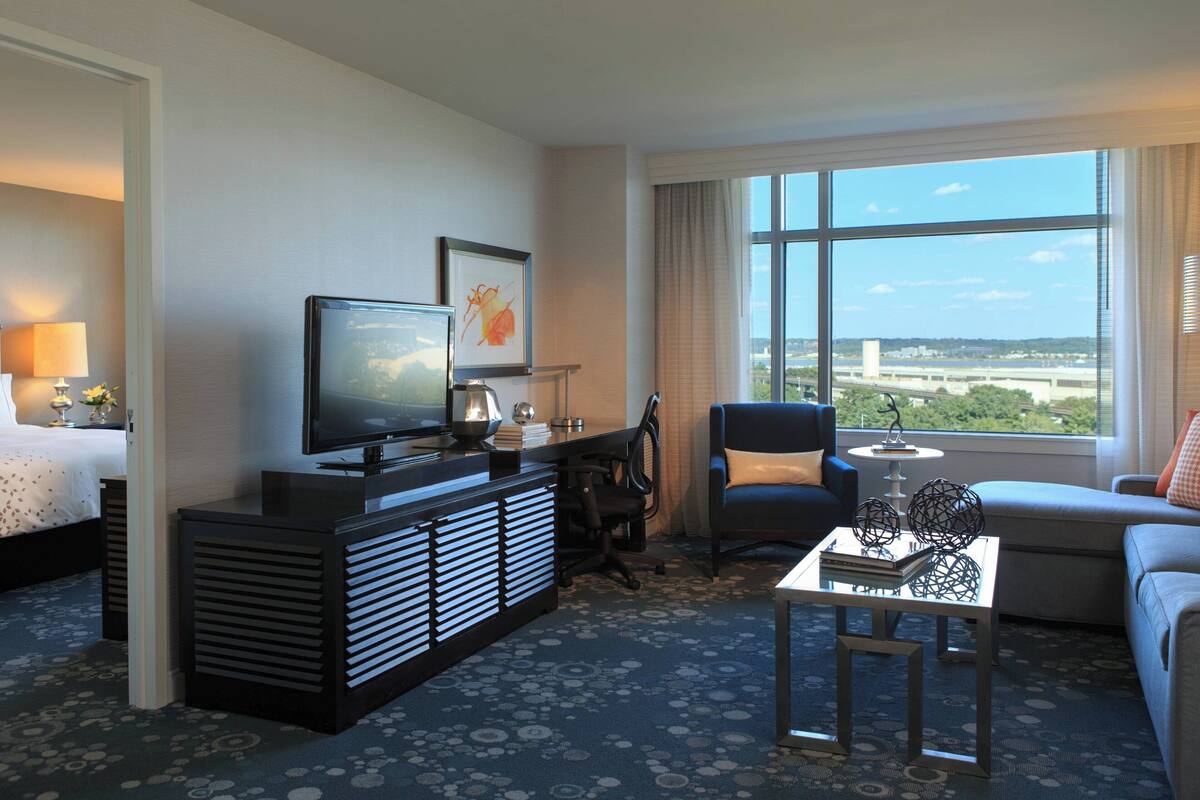 A hotel room with a bed, a desk with a TV, a chair, a sofa, and a large window offering a city view.
