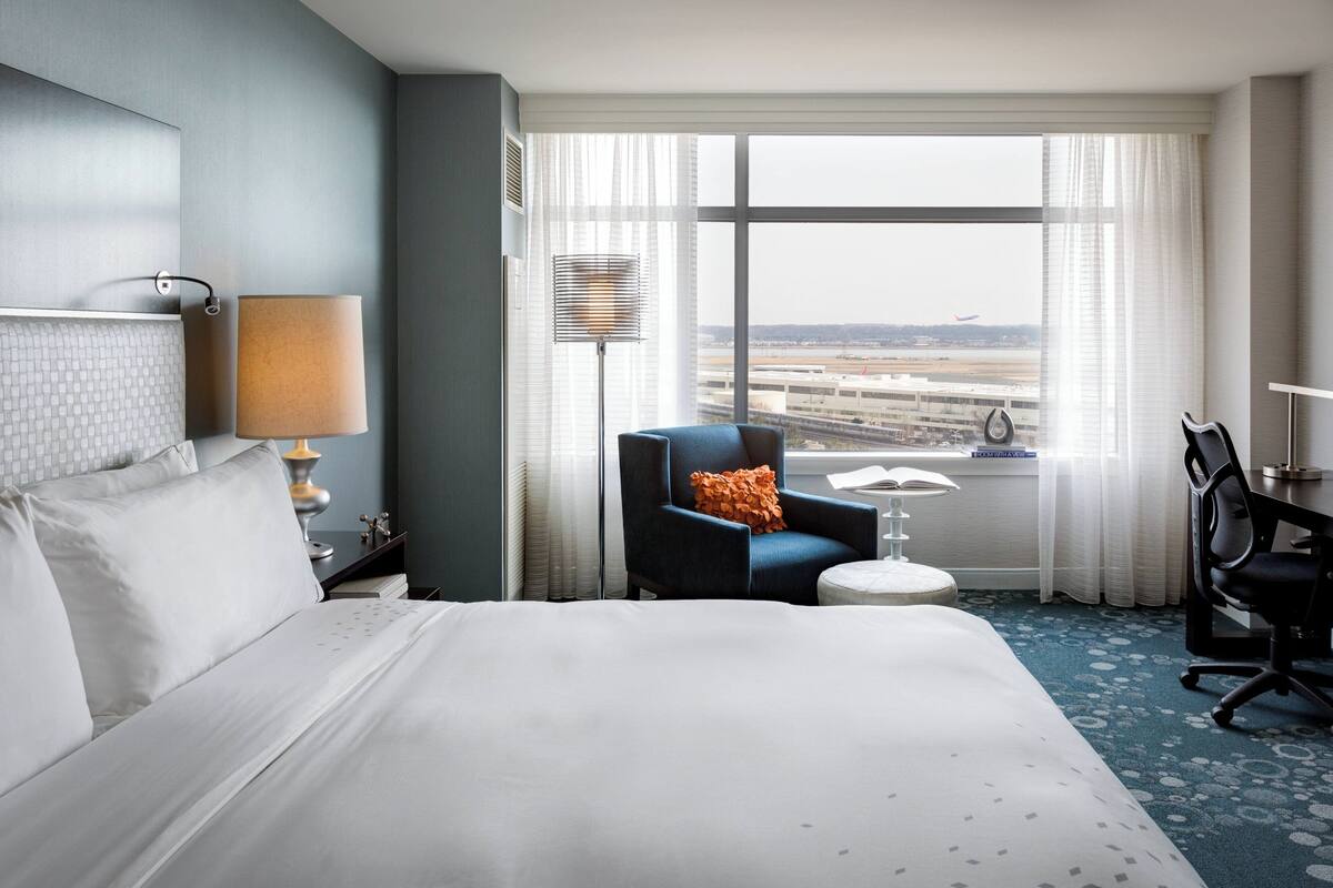 A hotel room with a large bed, a desk, a chair, a lamp, and a view of an airport.