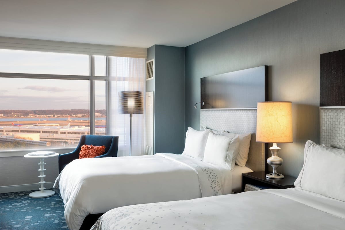 A hotel room with two beds, a view of the airport, and a small side table with a lamp.