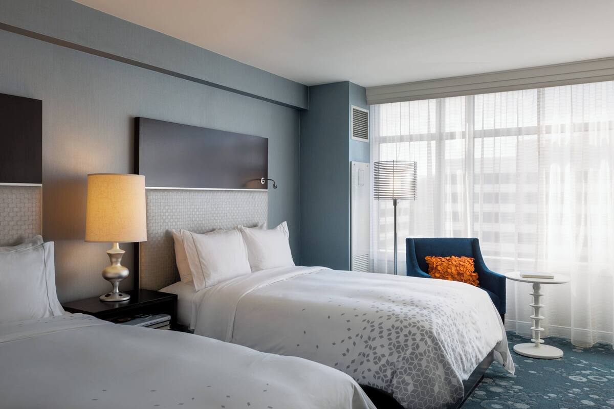 A hotel room with two beds, a bedside table with a lamp, a chair, a small round table, and a large window with sheer curtains.