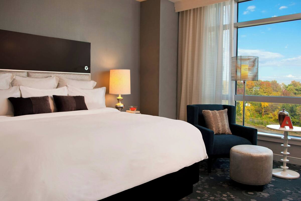 A hotel room with a large bed, a chair, an ottoman, a lamp, and a view of trees through a window.