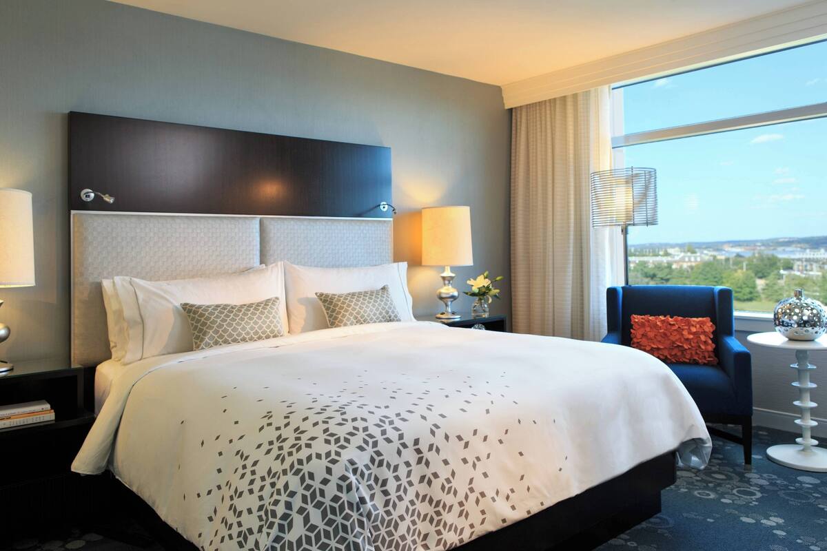 A hotel room with a large bed, a nightstand with a lamp, a chair, and a view of the city through the window.