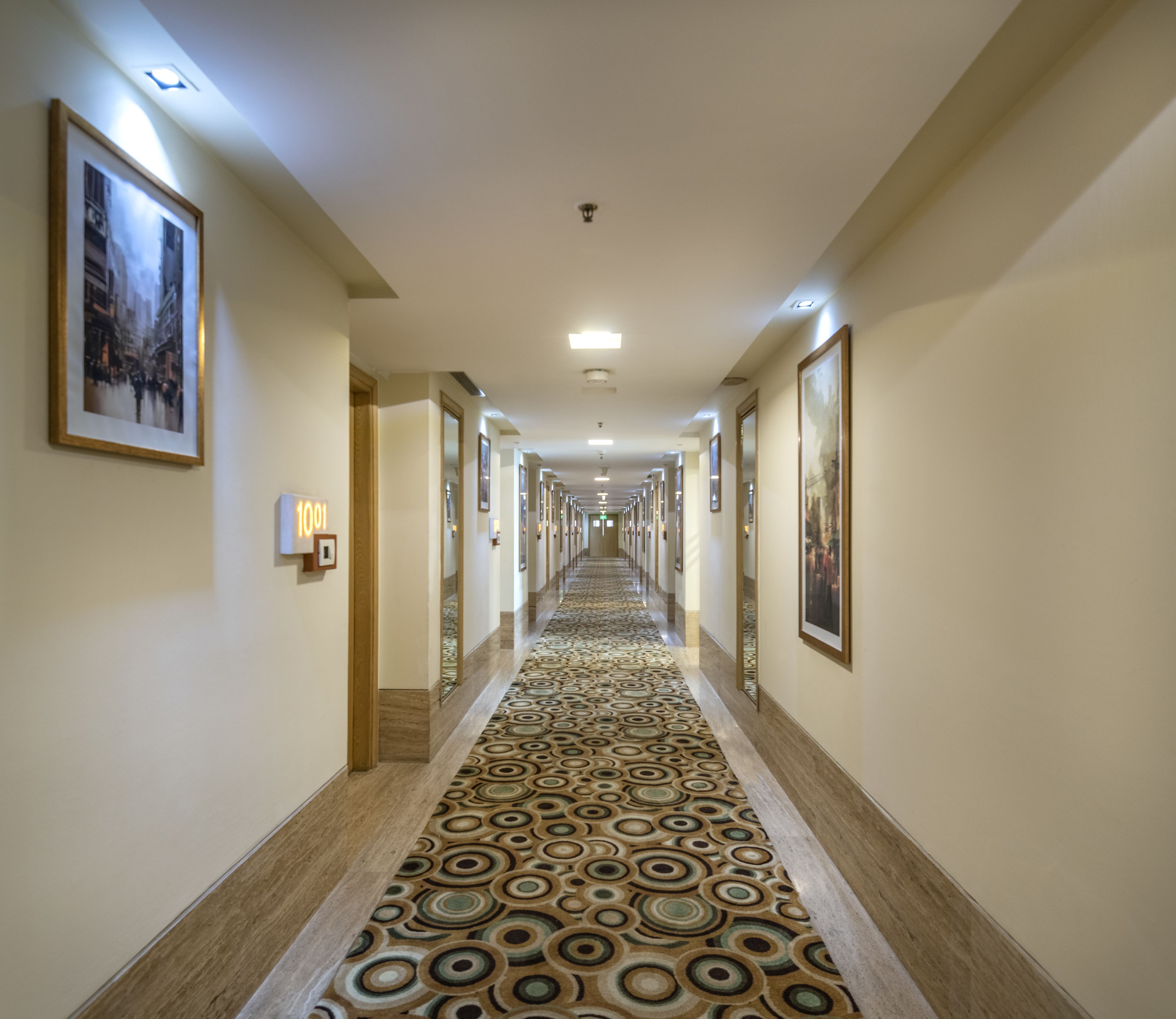 Een hotelgang met een patroon-tapijt, ingelijste foto's aan de muur, en een bordje met het kamernummer 1001.