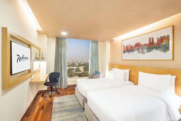 Una habitación de hotel con dos camas, un escritorio, una silla, un cuadro de paisaje urbano y un cuadro en la pared.