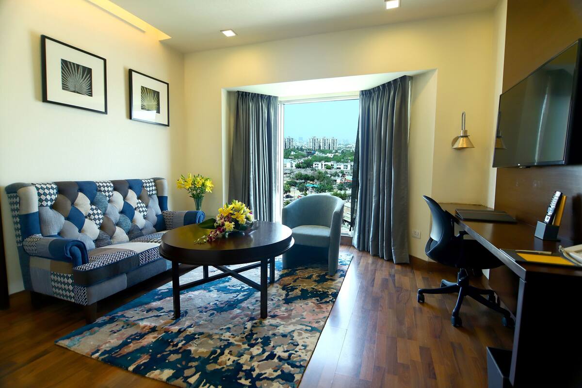 Sala de estar com sofá, mesa de centro redonda com flores, poltrona, escrivaninha com cadeira e televisão.