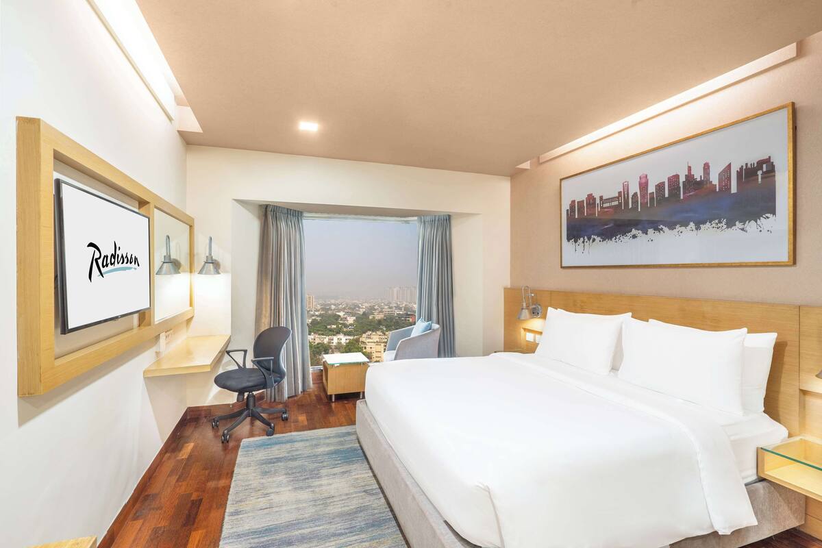 Quarto de hotel com uma cama grande, uma escrivaninha com cadeira, vista para a cidade e um quadro na parede.